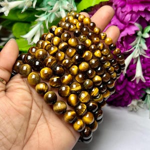 May include: A stack of tiger's eye gemstone bracelets with round beads. The bracelets are made of polished, golden brown stones with a shimmering, chatoyant effect.