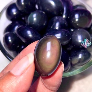 May include: A close-up of a hand holding a smooth, dark, iridescent obsidian egg. The egg is surrounded by other obsidian eggs in a clear bowl.