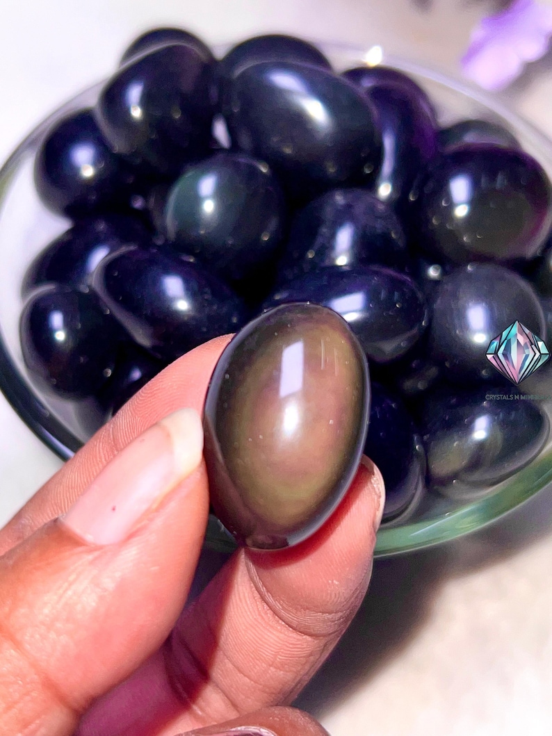 May include: A close-up of a hand holding a smooth, dark, iridescent obsidian egg. The egg is surrounded by other obsidian eggs in a clear bowl.