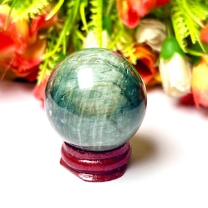 May include: A polished, green and blue crystal sphere sits on a small, round, dark red wooden stand. The sphere has a marbled appearance. The background features a blurred arrangement of red, orange, and white flowers with green foliage.