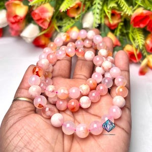 Puede incluir: Tres pulseras de piedras preciosas rosas con cuentas de diferentes tonos de rosa y naranja. Las pulseras se sostienen en una mano, con un fondo floral borroso. Las cuentas tienen aproximadamente 0,8 cm de diámetro.