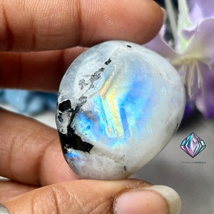 May include: A white moonstone with black inclusions and iridescent blue and gold flashes. The stone is smooth and polished, and it is held in a hand.