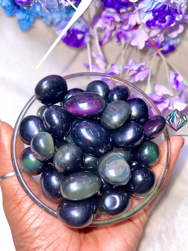 May include: A bowl of tumbled obsidian stones with iridescent green and purple hues. The stones are smooth and polished, and they have a dark, almost black, color.