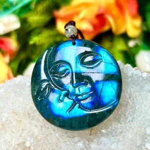 May include: A round, blue-green pendant with a carved face of the sun and moon. The pendant is made of a stone with a shimmering, iridescent quality. It hangs from a brown cord with a small, round bead. The background is blurred with colorful flowers.