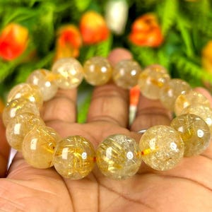 May include: A golden-yellow beaded bracelet with inclusions, held in a hand. The beads are round and translucent, with golden strands visible inside. The bracelet is set against a blurred background of green and orange.