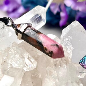 May include: A pink and black crystal pendant on a black cord. The pendant is a pointed, faceted crystal with a silver setting. The crystal is resting on a bed of white crystals.