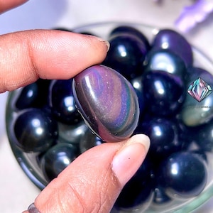 May include: A close-up of a collection of iridescent black obsidian tumbled stones. The stones are smooth and polished, with a deep black color and a subtle iridescent sheen.