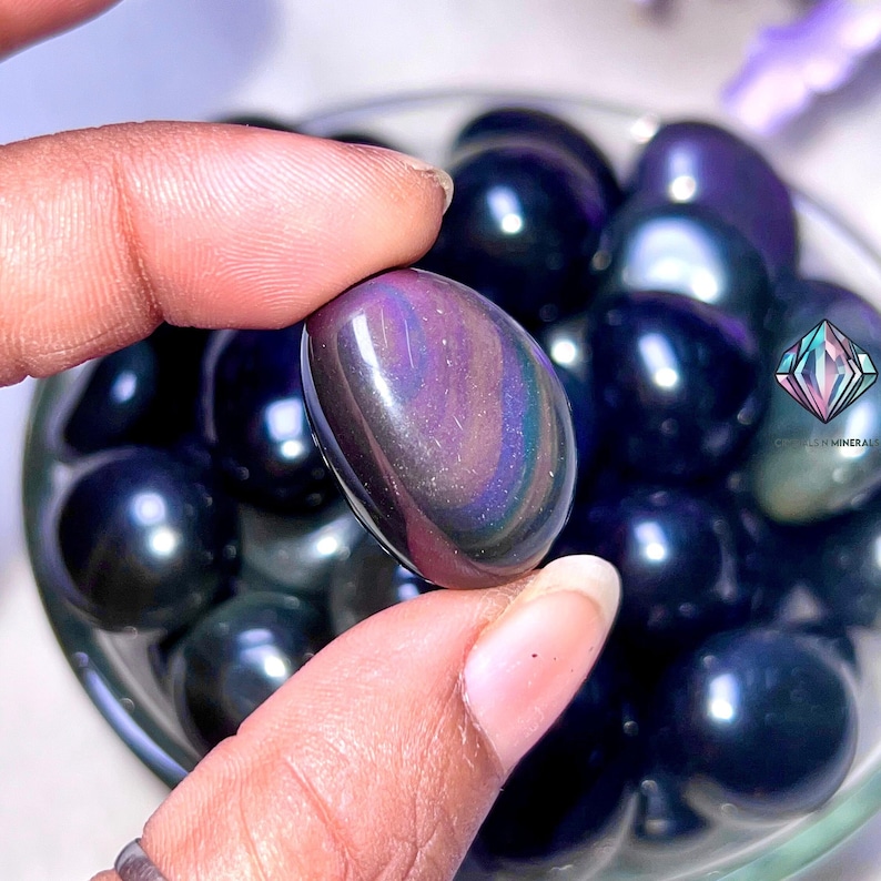 May include: A close-up of a collection of iridescent black obsidian tumbled stones. The stones are smooth and polished, with a deep black color and a subtle iridescent sheen.