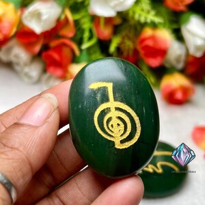 May include: Green stone with a gold-coloured symbol, known as the "Sacred Geometry" symbol, on it. The stone is smooth and oval-shaped.