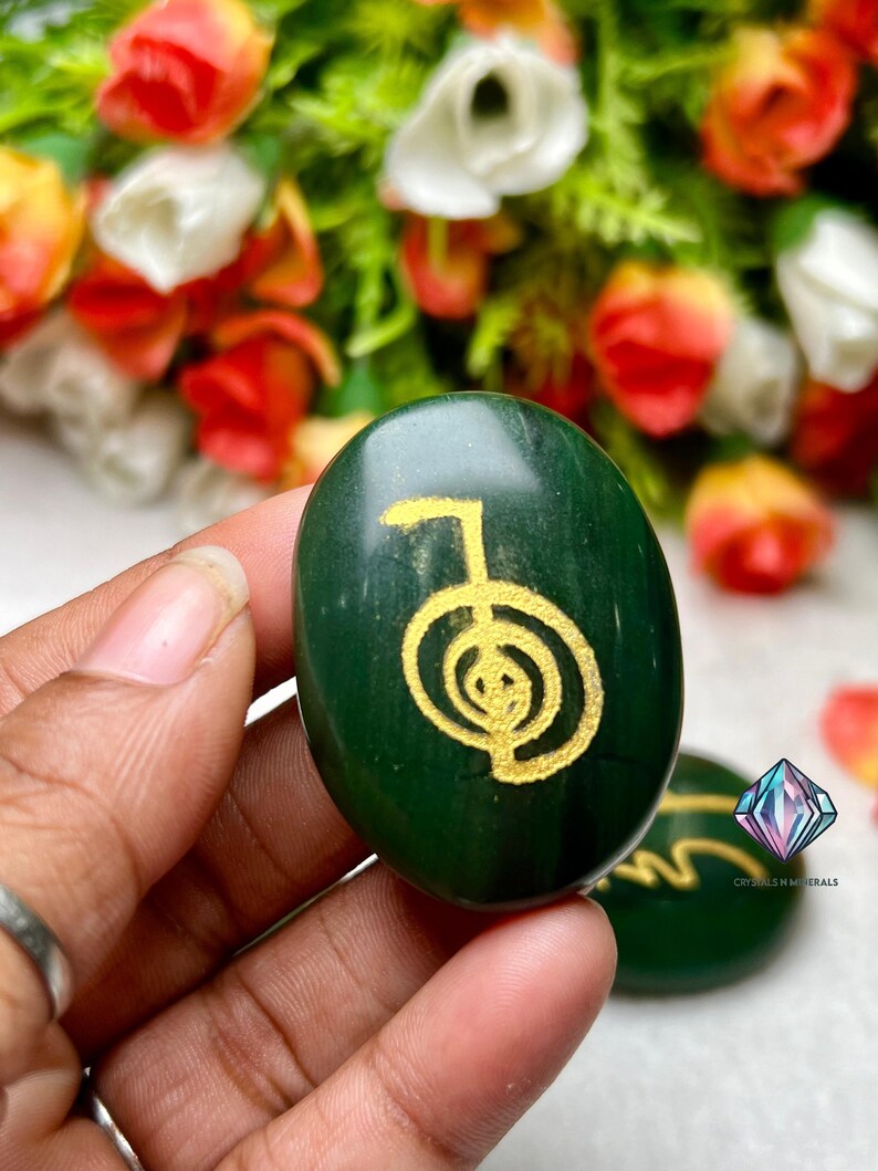 May include: Green stone with a gold-coloured symbol, known as the "Sacred Geometry" symbol, on it. The stone is smooth and oval-shaped.