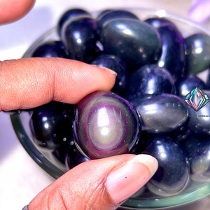 May include: A close-up of a bowl filled with iridescent black obsidian tumbled stones. The stones have a rainbow sheen and are smooth and polished.