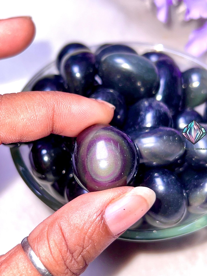 May include: A close-up of a bowl filled with iridescent black obsidian tumbled stones. The stones have a rainbow sheen and are smooth and polished.