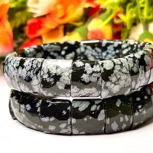 Puede incluir: Dos pulseras de obsidiana copo de nieve negras y blancas. Las pulseras están hechas de cuentas cuadradas con un patrón moteado.