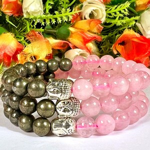 May include: Two beaded bracelets. One bracelet features dark gray pyrite beads and silver Buddha head beads. The other bracelet has pink rose quartz beads. The bracelets are displayed in front of a colorful floral arrangement.