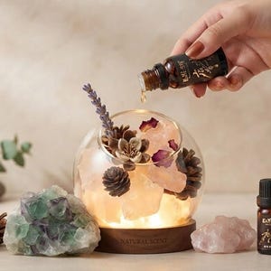 Puede incluir: Un difusor de cristal sobre base de madera con cristales rosas, piñas, pétalos de rosa secos y una rama de lavanda. Un frasco marrón con tapón negro se sostiene encima, goteando aceite en el cuenco. Un grupo de fluorita se sitúa al lado.