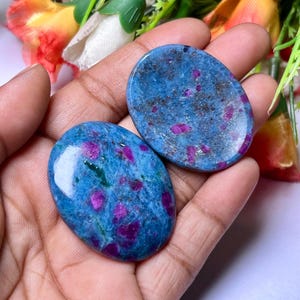 May include: Two oval-shaped blue kyanite stones with pink ruby inclusions. The stones are polished and have a smooth, glossy finish.
