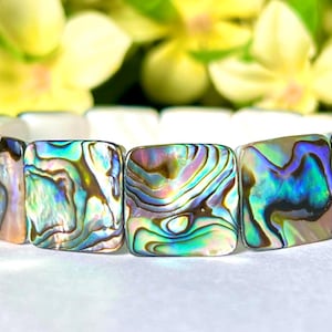May include: Bracelet made from abalone shell, displaying iridescent hues of blue, green, and purple. The square-shaped abalone beads are strung together. The background features blurred yellow flowers and green leaves.