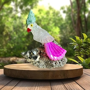 May include: A clear glass parrot sculpture with a teal crest and a bright pink tail, perched on a textured rock. The bird has a red beak and yellow eyes. The sculpture is displayed on a wooden platform.