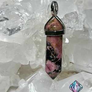 May include: A pink and black rhodochrosite crystal pendant with a silver setting. The pendant is shaped like a point and is surrounded by white crystal rocks.