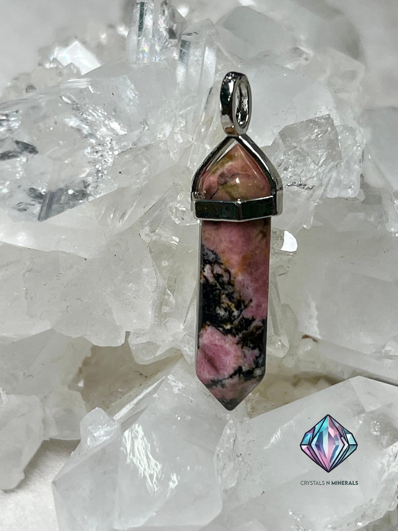 May include: A pink and black rhodochrosite crystal pendant with a silver setting. The pendant is shaped like a point and is surrounded by white crystal rocks.