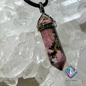 May include: A pink and black rhodochrosite crystal pendant with a silver setting. The pendant is hanging from a black cord and is resting on a bed of clear quartz crystals.