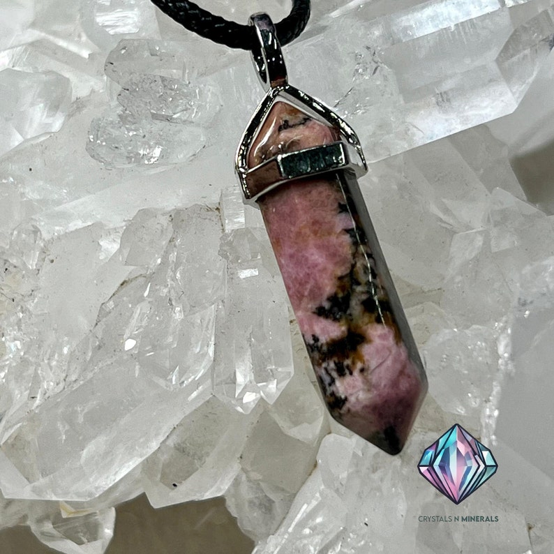 May include: A pink and black rhodochrosite crystal pendant with a silver setting. The pendant is hanging from a black cord and is resting on a bed of clear quartz crystals.