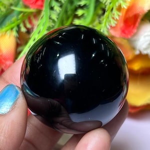 Könnte beinhalten: Eine glatte, schwarze Obsidiankugel, poliert auf einen hohen Glanz. Die Kugel wird in einer Hand gehalten und reflektiert das Licht, wodurch eine dunkle, spiegelartige Oberfläche entsteht.