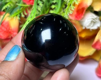 May include: A smooth, black obsidian sphere, polished to a high shine. The sphere is held in a hand, reflecting light and creating a dark, mirror-like surface.