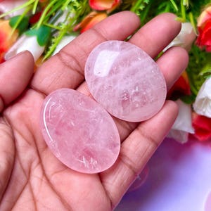 May include: Two oval-shaped rose quartz stones, one on top of the other, in a hand. The stones are a pale pink color and have a smooth, polished surface.