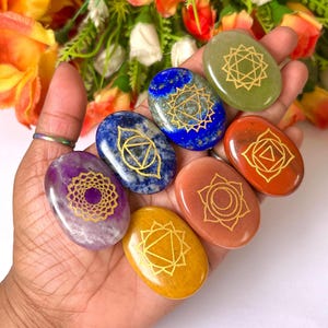 May include: Seven smooth, oval-shaped stones with gold chakra symbols. The stones are arranged in a rainbow pattern, from purple to red.