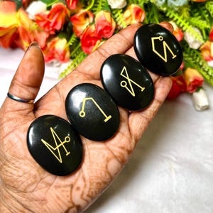 May include: Five black oval-shaped stones with gold symbols etched on them. The stones are held in a hand.