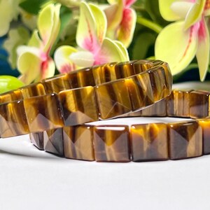 Peut inclure: Deux bracelets en œil de tigre brun et or avec des perles de forme carrée. Les bracelets sont empilés l'un sur l'autre.