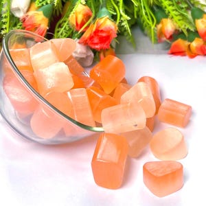 May include: A glass bowl filled with orange and pink heart-shaped selenite crystals. The bowl is tipped over, spilling some of the crystals onto a white surface.