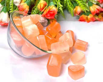 May include: A glass bowl filled with orange and pink heart-shaped selenite crystals. The bowl is tipped over, spilling some of the crystals onto a white surface.