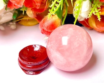 May include: A pink rose quartz sphere on a red wooden stand. The sphere is smooth and polished, with a slight shimmer. The stand is made of wood and has a natural finish.