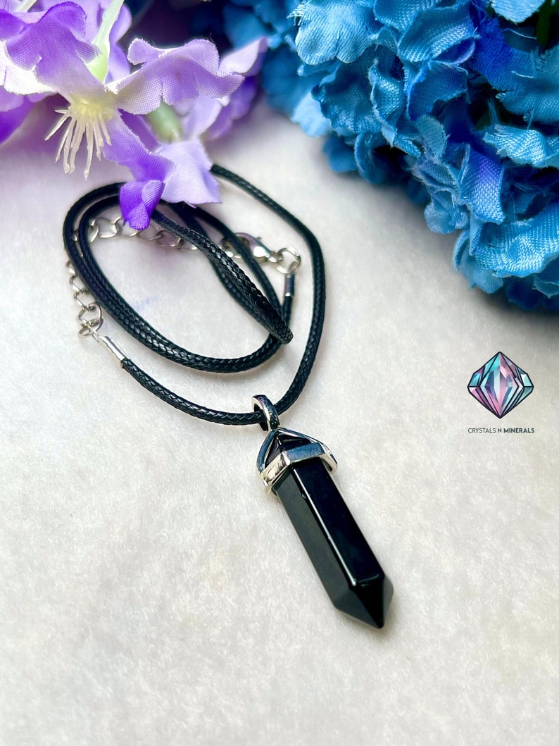 Puede incluir: Collar con colgante de punta de obsidiana negra con engaste plateado en un cord&oacute;n negro. Crystals N Minerals est&aacute; impreso en la imagen.