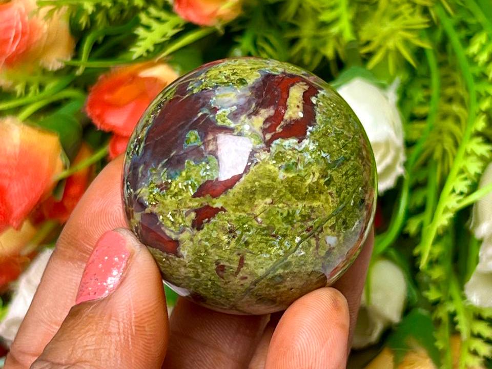 May include: Green and red jasper sphere, polished and smooth, with swirling patterns of color. The sphere is held in a hand.