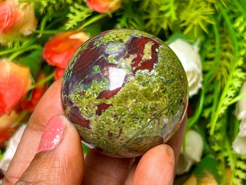 May include: Green and red jasper sphere, polished and smooth, with swirling patterns of color. The sphere is held in a hand.