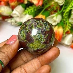 May include: A green, red, and brown sphere with a speckled pattern. The sphere is made of polished stone and is about the size of a golf ball.