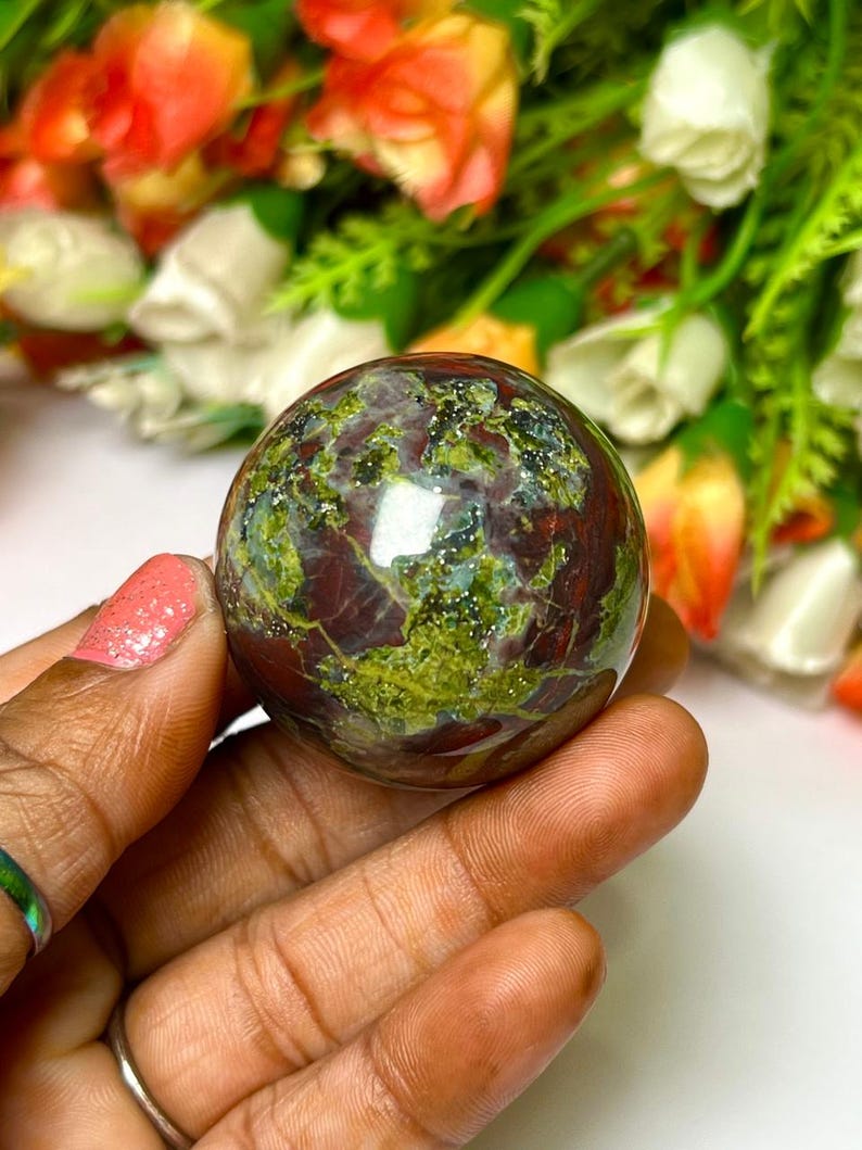 May include: A green, red, and brown sphere with a speckled pattern. The sphere is made of polished stone and is about the size of a golf ball.