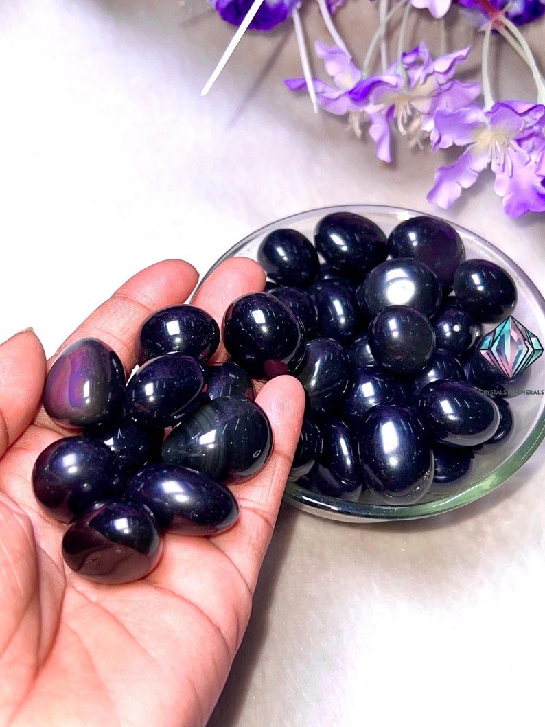 May include: A collection of black obsidian tumbled stones, also known as black glass, in a small bowl. The stones are smooth and polished, with a shiny, iridescent finish.
