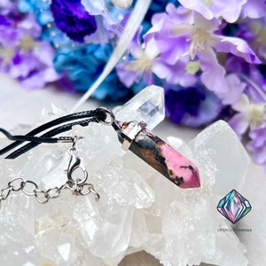 May include: A pink and black crystal pendant on a black cord necklace with a silver chain clasp. The pendant is resting on a bed of white crystals. The text "CRYSTALS & MINERALS" is visible in the bottom right corner of the image.