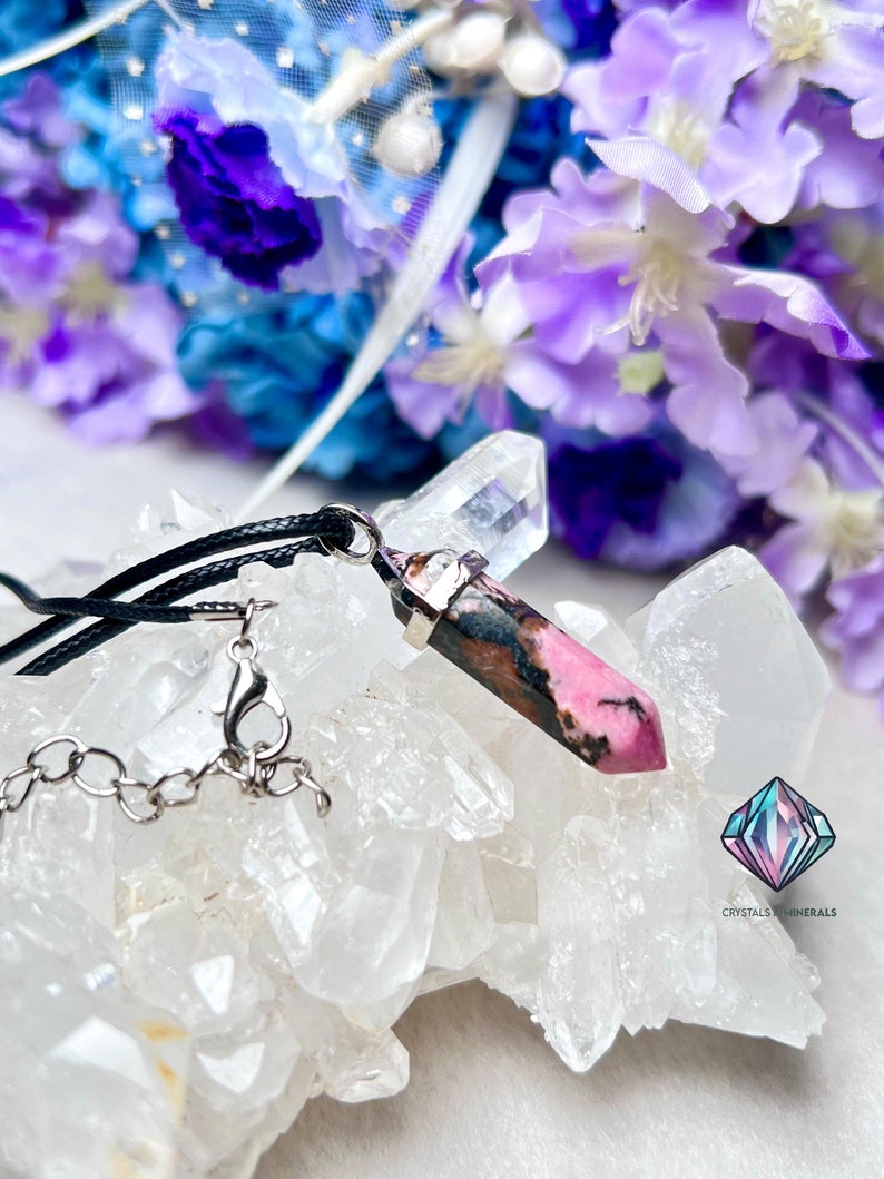 May include: A pink and black crystal pendant on a black cord necklace with a silver chain clasp. The pendant is resting on a bed of white crystals. The text "CRYSTALS & MINERALS" is visible in the bottom right corner of the image.