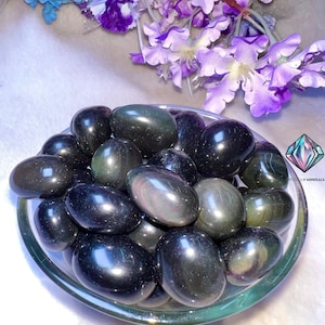 May include: A bowl of black obsidian tumbled stones with iridescent rainbow flashes. The stones are smooth and polished, and they are arranged in a random pattern. The bowl is clear glass and has a white background.