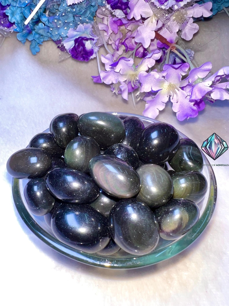 May include: A bowl of black obsidian tumbled stones with iridescent rainbow flashes. The stones are smooth and polished, and they are arranged in a random pattern. The bowl is clear glass and has a white background.