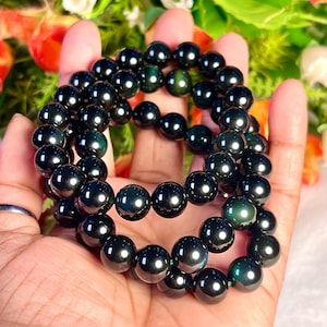 May include: A black beaded bracelet with iridescent green and blue hues. The beads are round and smooth, and the bracelet is layered.
