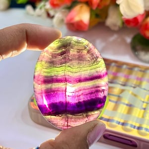 May include: A smooth, oval-shaped fluorite stone with vibrant green, purple, and pink banding. The stone is polished and has a translucent appearance.