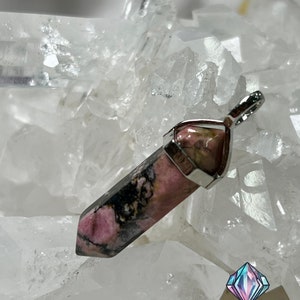 May include: A pink and black gemstone pendant with a silver setting. The pendant is shaped like a point and is set on a bed of white crystals.