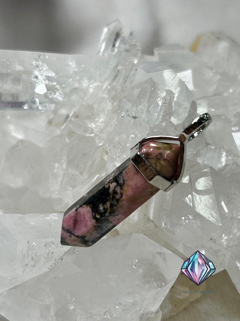 May include: A pink and black gemstone pendant with a silver setting. The pendant is shaped like a point and is set on a bed of white crystals.