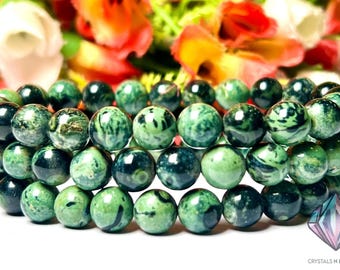 May include: A stack of three bracelets made of round, green beads with dark green and black marbling. The beads are arranged in rows, and the background features blurred flowers in shades of orange, white, and yellow.